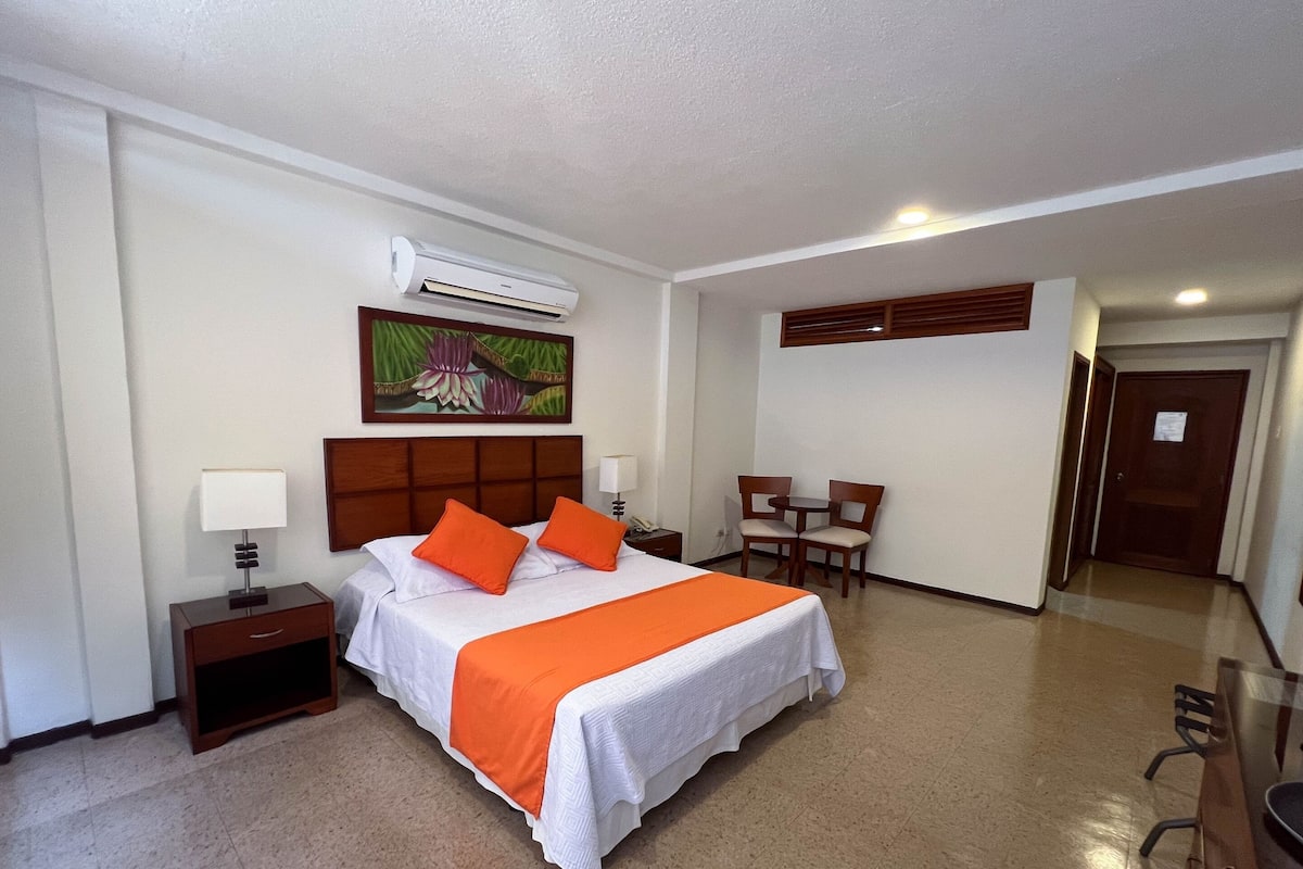A hotel room with a bed, a desk, a chair, a nightstand, a lamp, and an air conditioning unit.
