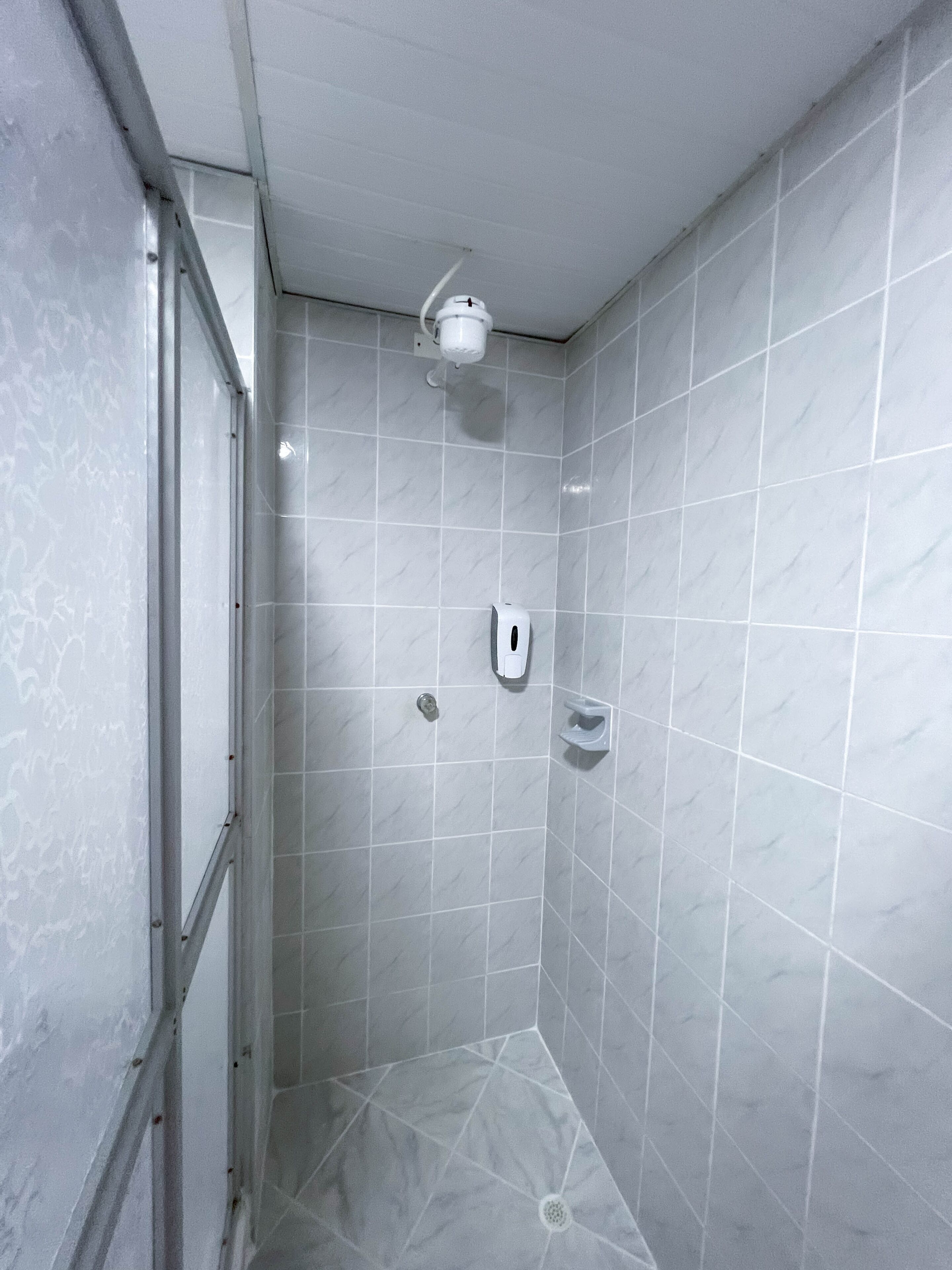 Un baño pequeño con azulejos, una ducha de techo y un dispensador de jabón en la pared.