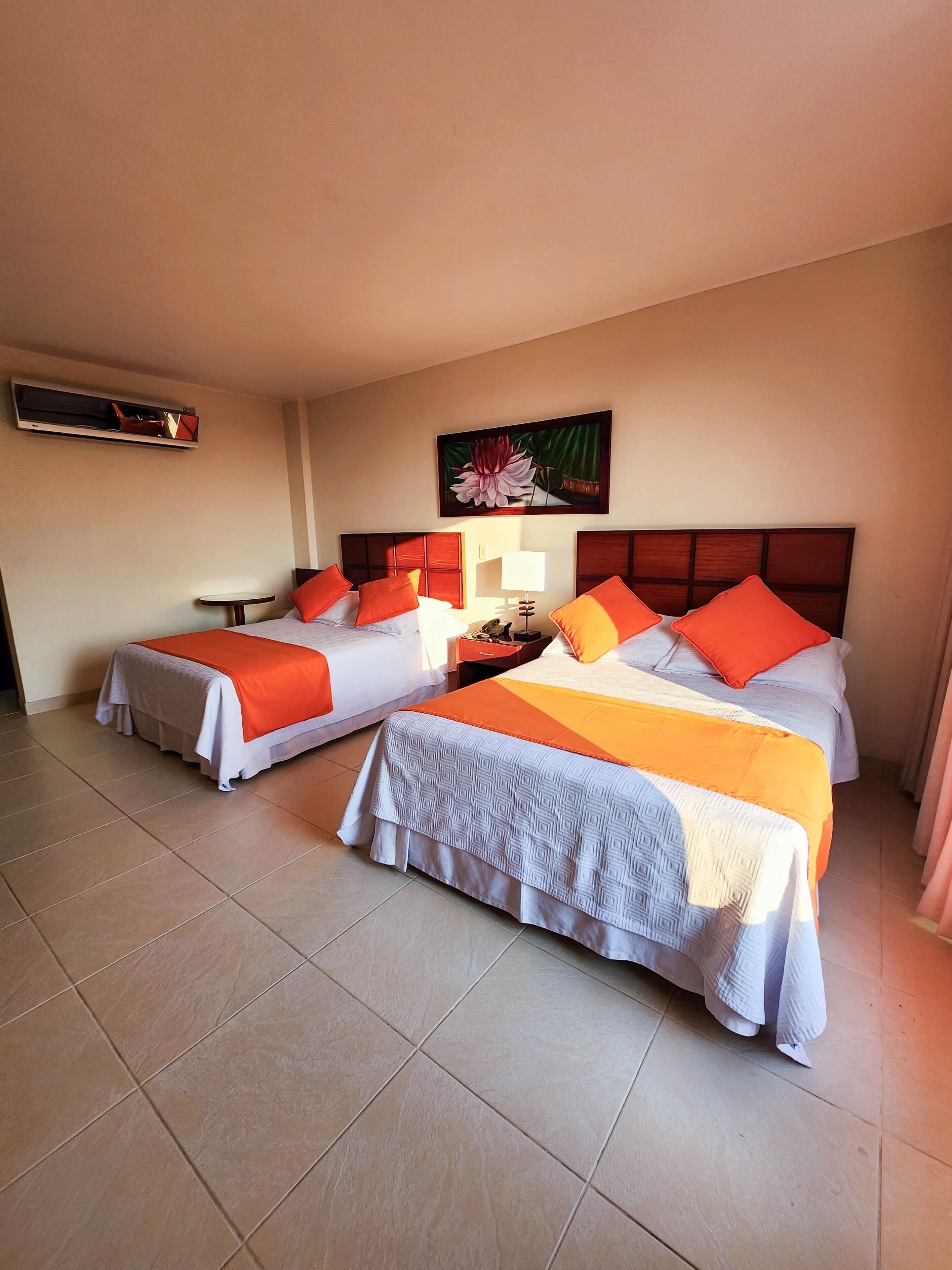 Habitación de hotel con dos camas, cada una con ropa y almohadas anaranjadas, una mesita de noche pequeña con una lámpara, y aire acondicionado instalado en la pared.