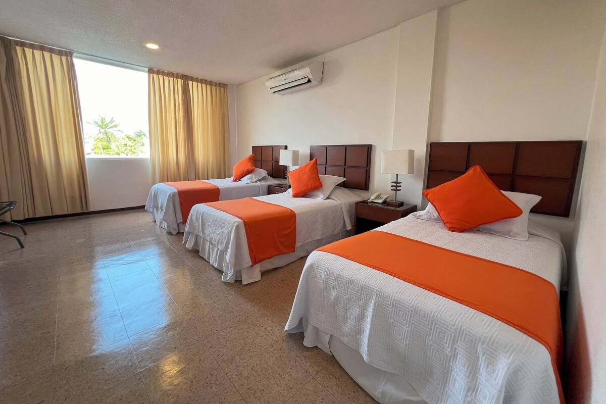 A hotel room with three beds, each with an orange bedspread and white pillows. There is a window with curtains, an air conditioning unit on the wall, and a tiled floor.