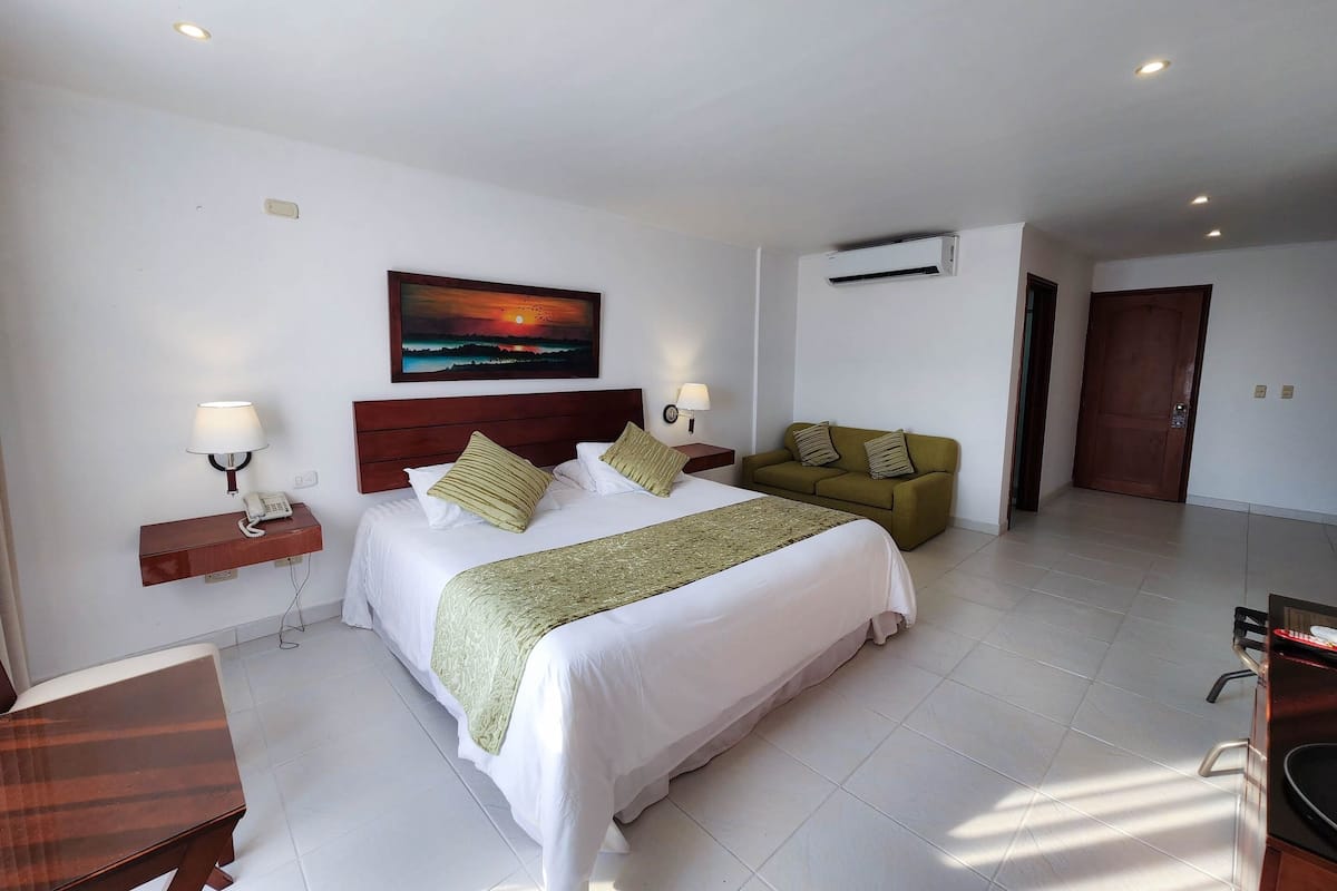 A hotel room with a large bed, a green sofa, a painting of a sunset, and a desk with a lamp.