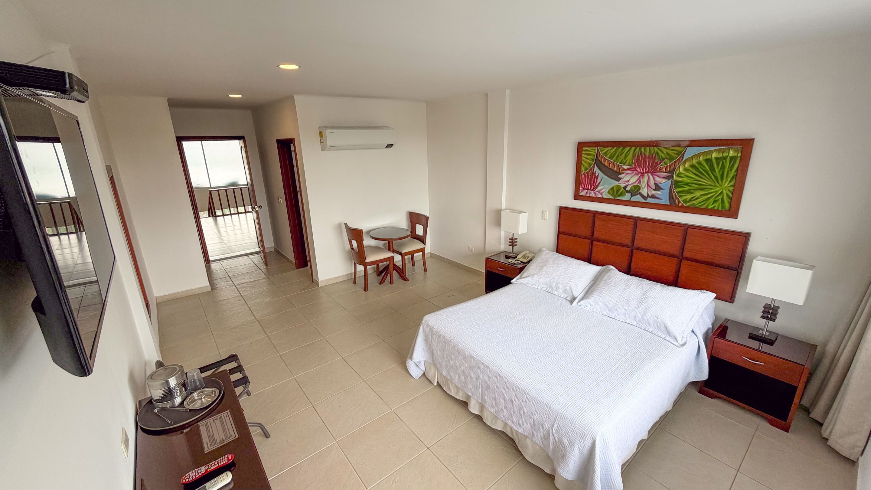 A hotel room with a bed, bedside table, chair, and a dining table with chairs. There is a TV, a painting on the wall, and an air conditioning unit.