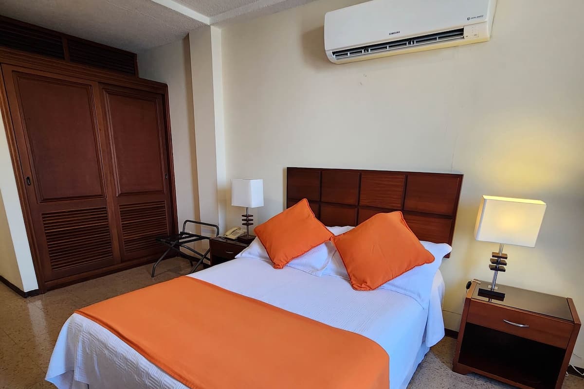 A hotel room with a bed, an air conditioning unit, a nightstand, and a wardrobe.