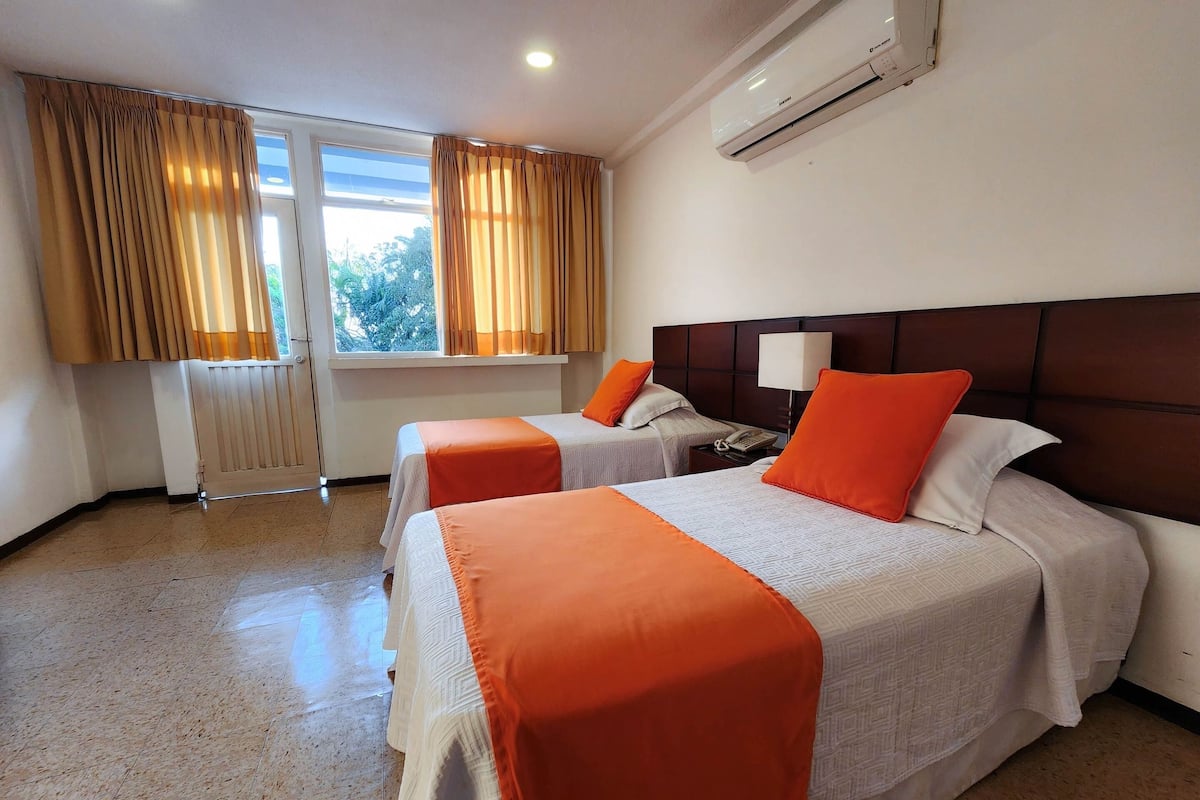 A hotel room with two beds, a window with curtains, an air conditioning unit, and a bedside table with a lamp.