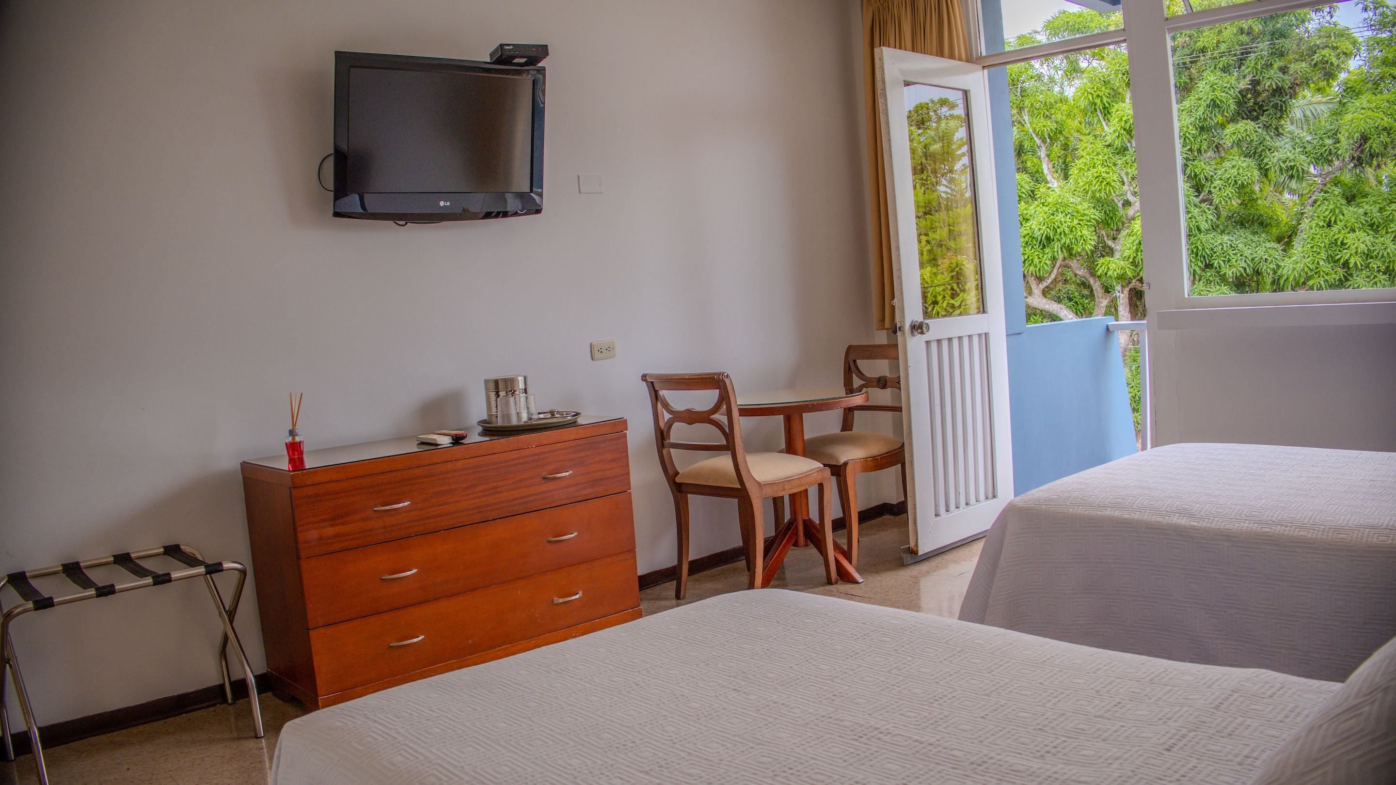 A hotel room with two beds, a wooden dresser, a television mounted on the wall, and a window with a view of greenery.