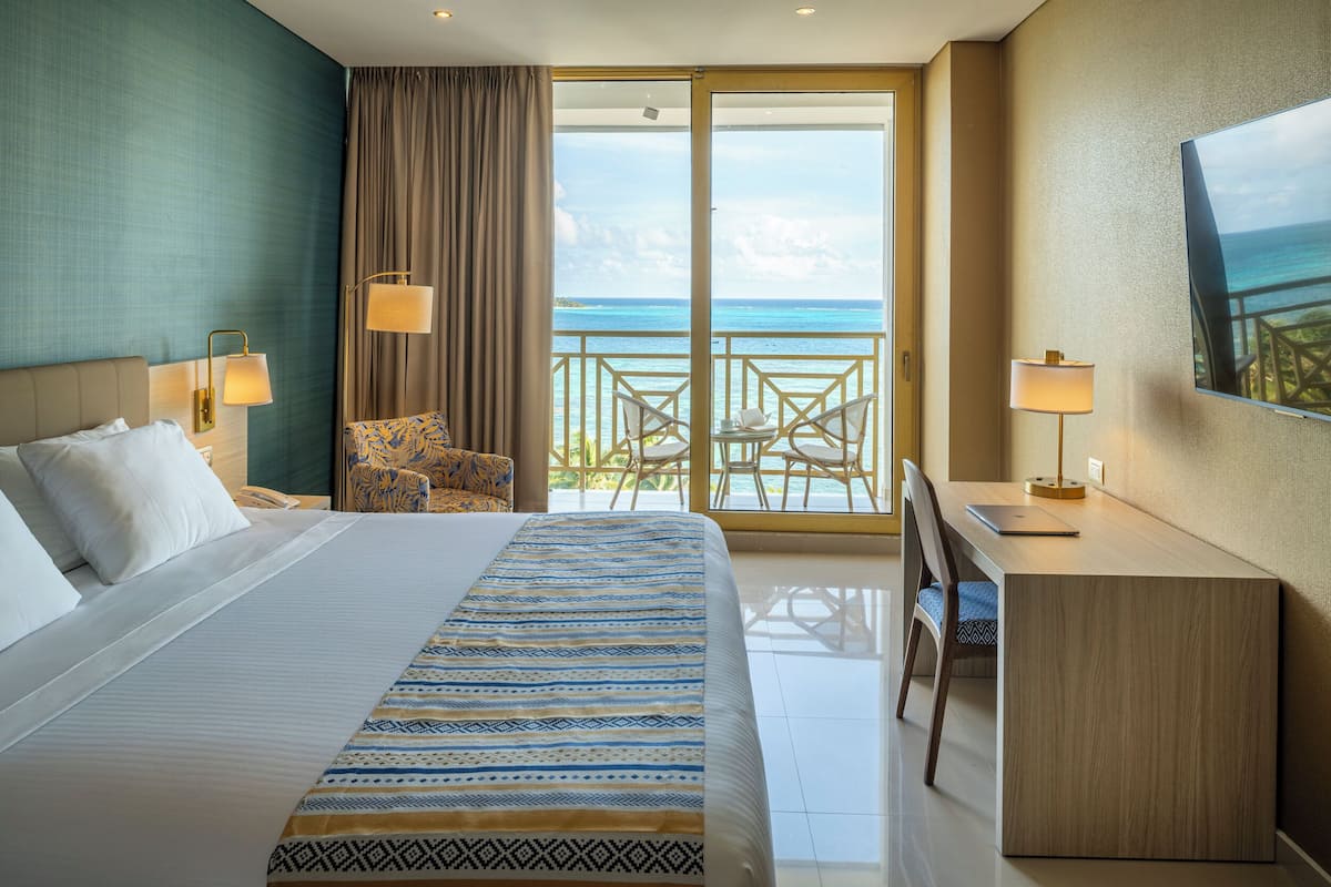 A hotel room with a bed, a desk, a chair, and a balcony with a view of the ocean.