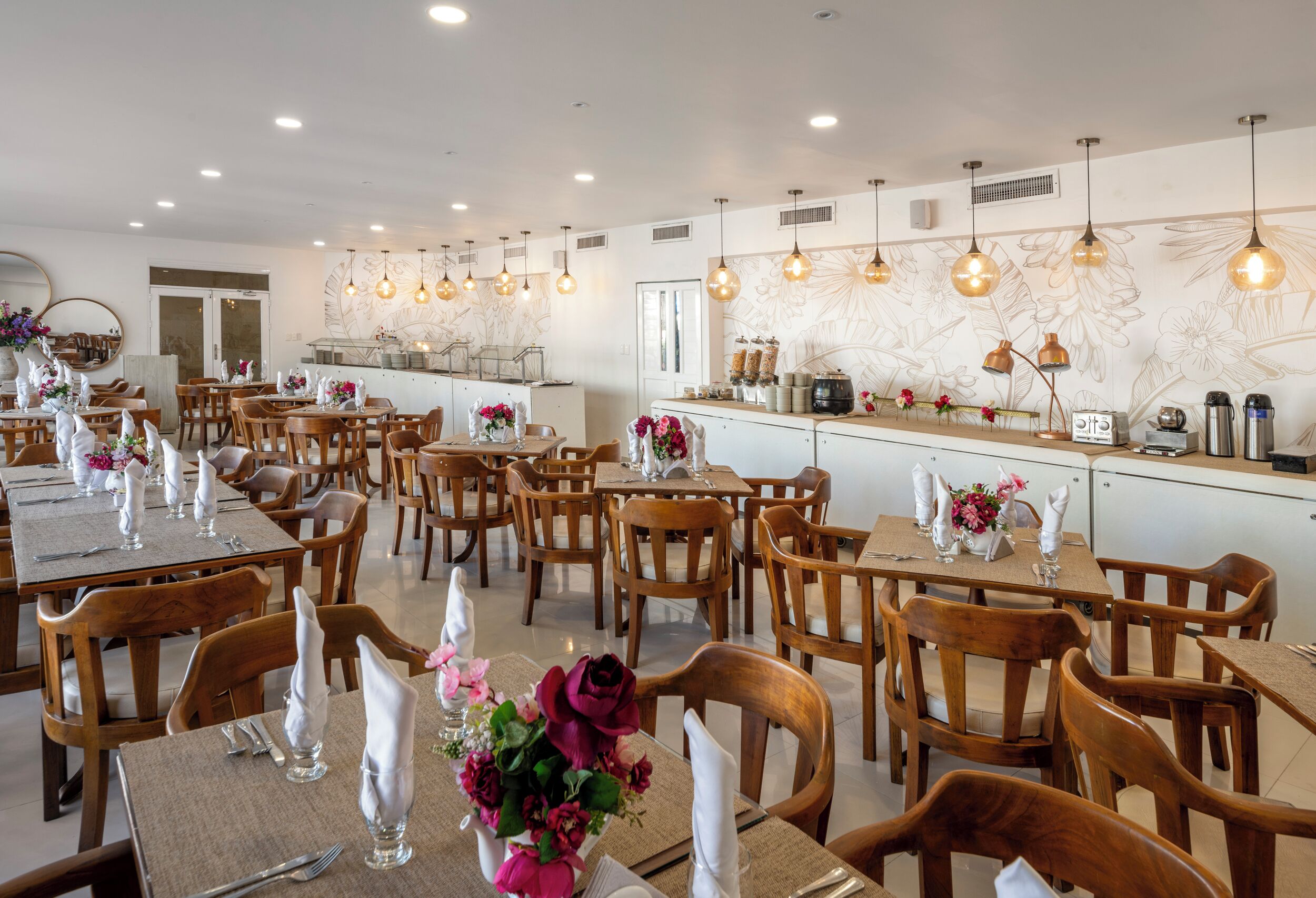 Un restaurante con mesas y sillas de madera, servilletas blancas y centros de mesa con flores.