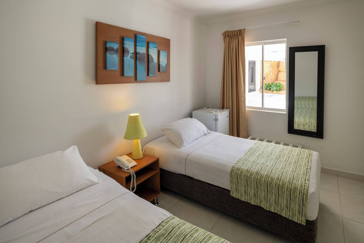 A hotel room with two beds, a bedside table with a lamp, a telephone, and a window with curtains.