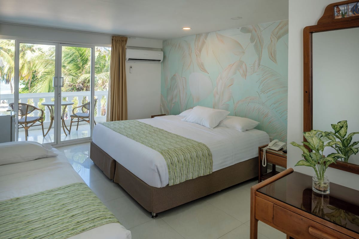 A hotel room with a large bed, a smaller bed, a desk, and a chair. There is a balcony with a table and chairs.