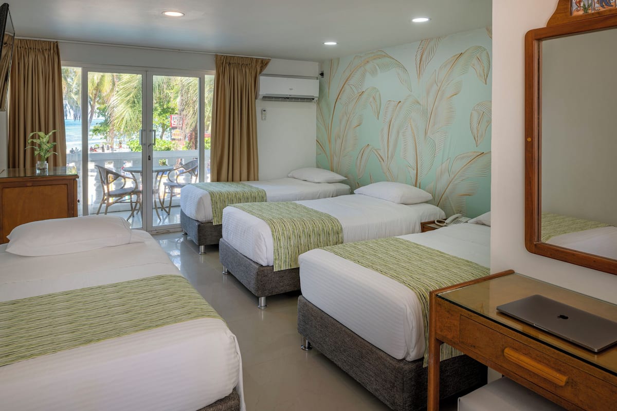 A hotel room with three beds, a glass desk with a laptop, and a balcony with a view of the sea.