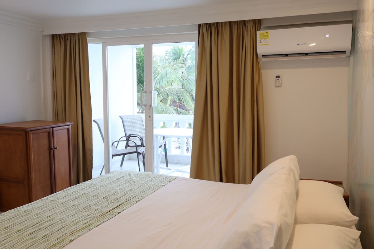 A hotel room with a bed, a wardrobe, an air conditioning unit, and a balcony with a chair and table.