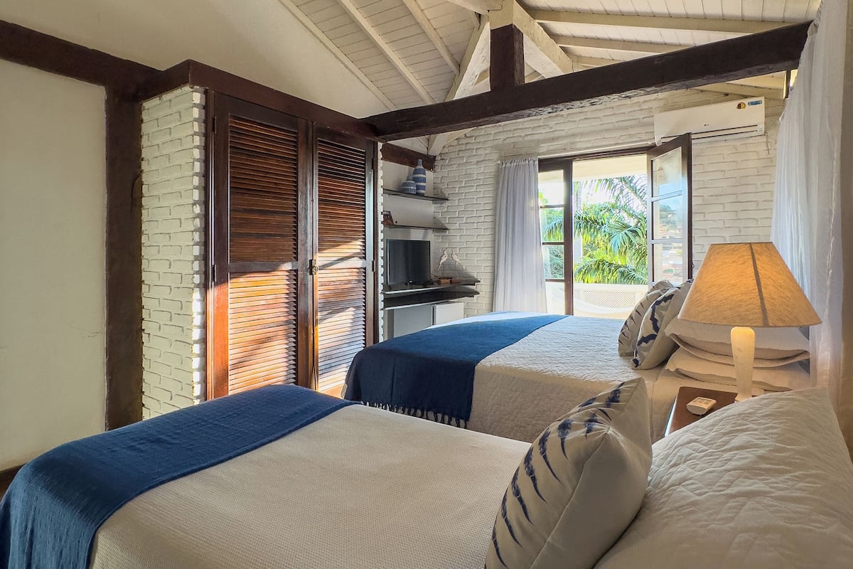 A room with two beds, a brick wall, wooden beams, a window with blinds, a lamp, and a shelf with decorative items.