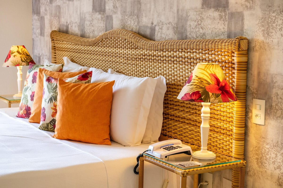 A bed with a woven headboard, white and orange pillows, a bedside lamp, and a telephone on a glass table.