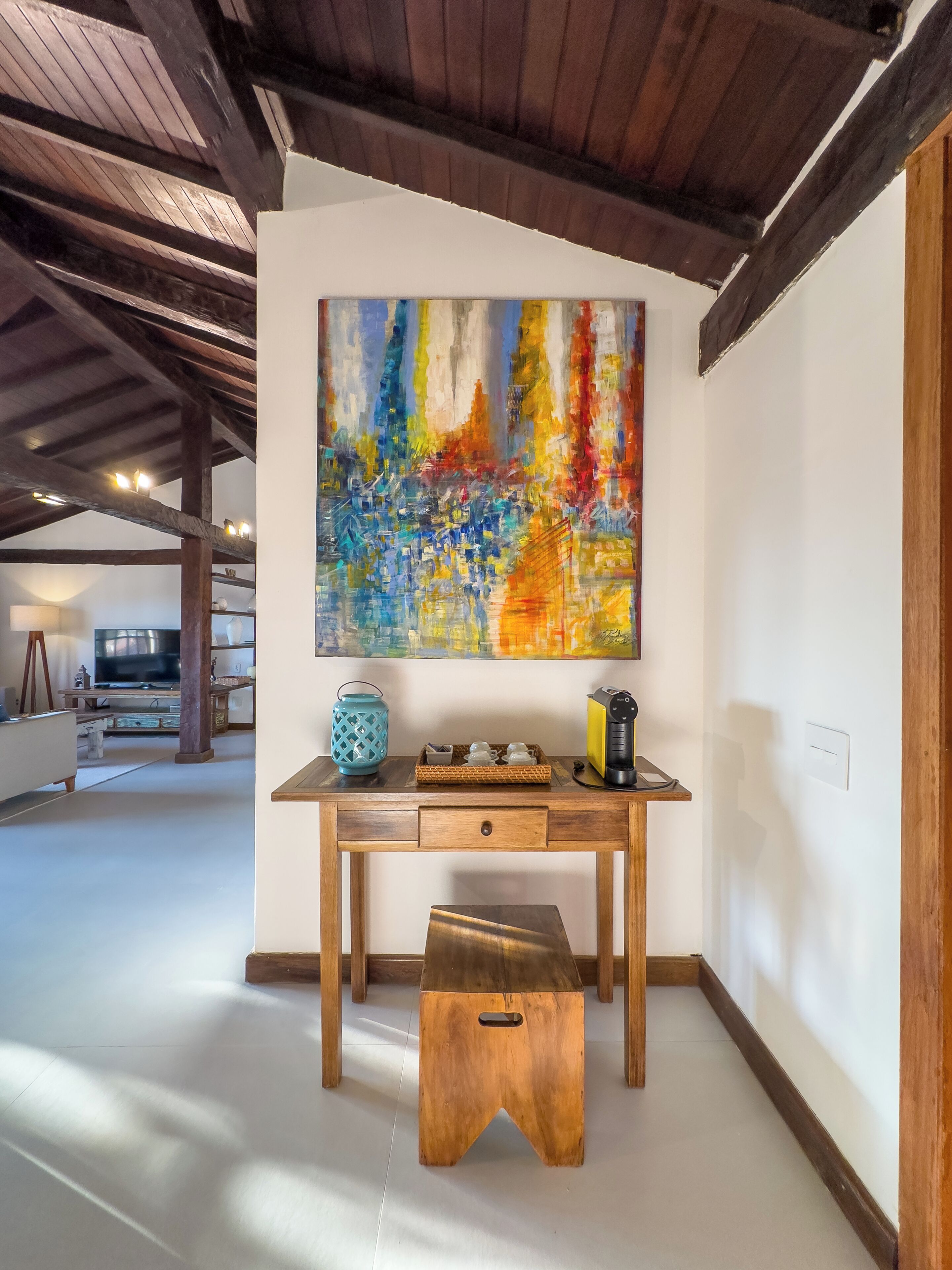 Una scrivania in legno con un quadro astratto dai colori vivaci sopra, un piccolo sgabello di fronte e una panca in legno sullo sfondo.