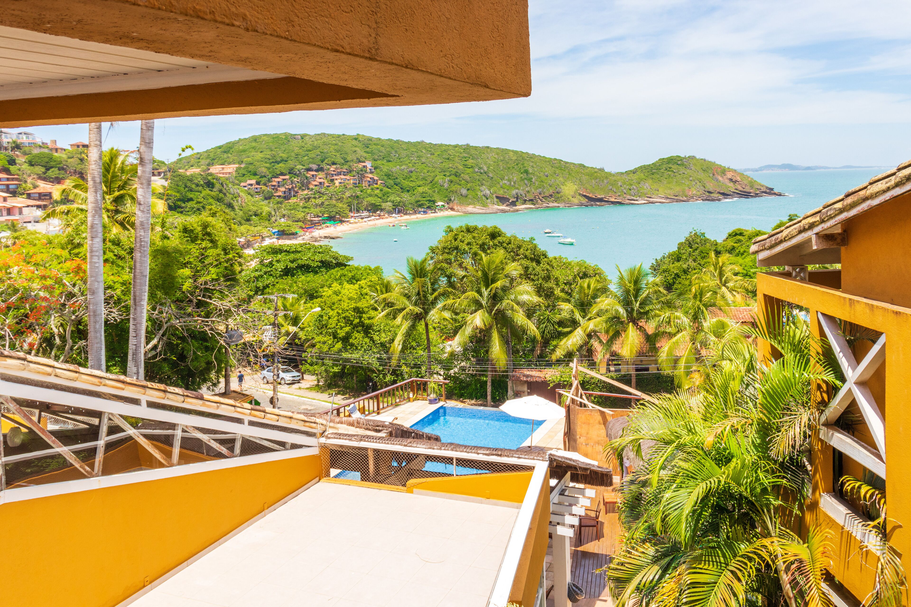 Une vue depuis un balcon donnant sur une plage, avec une piscine, des palmiers et un paysage vallonné en arrière-plan.