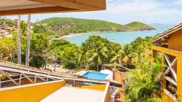 Une vue depuis un balcon donnant sur une plage, avec une piscine, des palmiers et un paysage vallonné en arrière-plan.