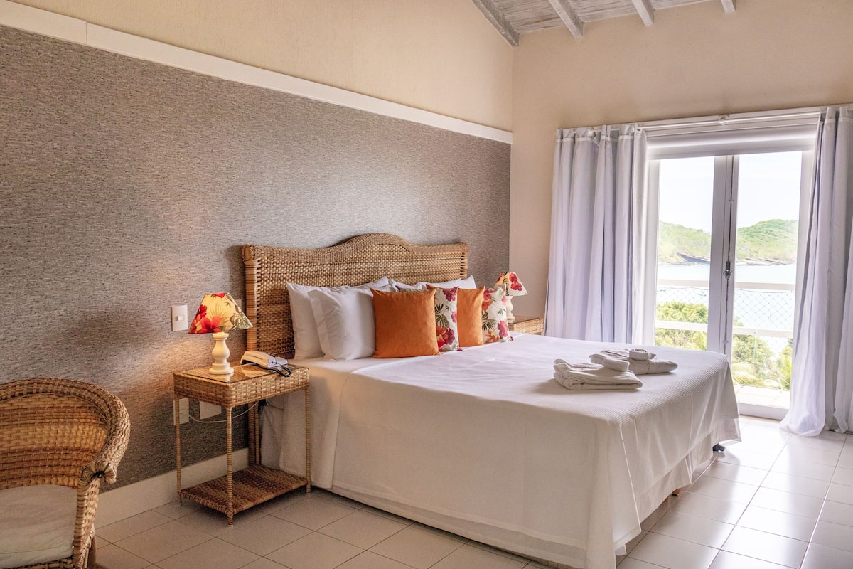 A bedroom with a large bed, a wicker chair, a nightstand with a lamp, and a view of the outdoors through a window.