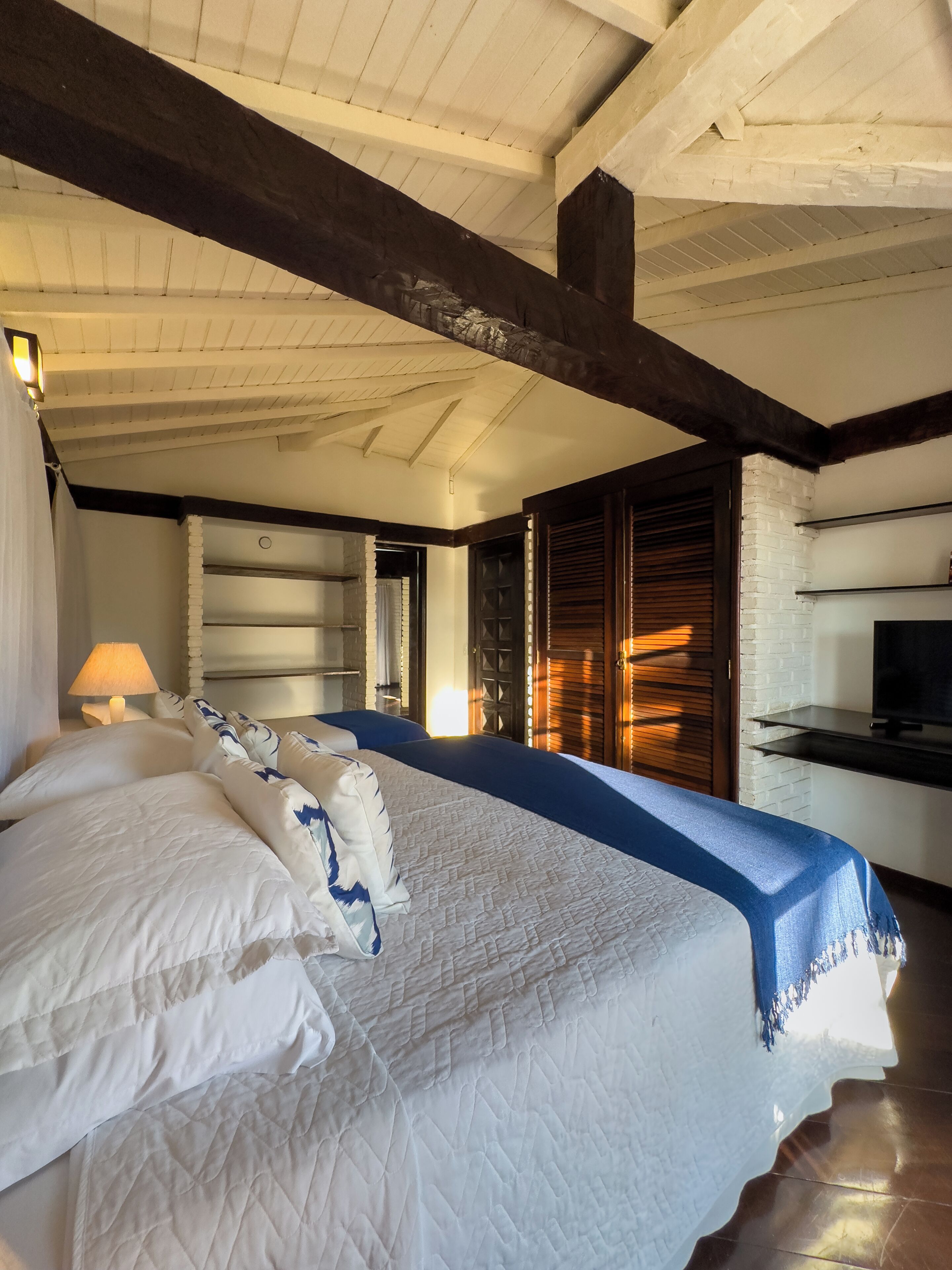 A bedroom with a large bed, a nightstand with a lamp, a TV on a shelf, and a brick wall.