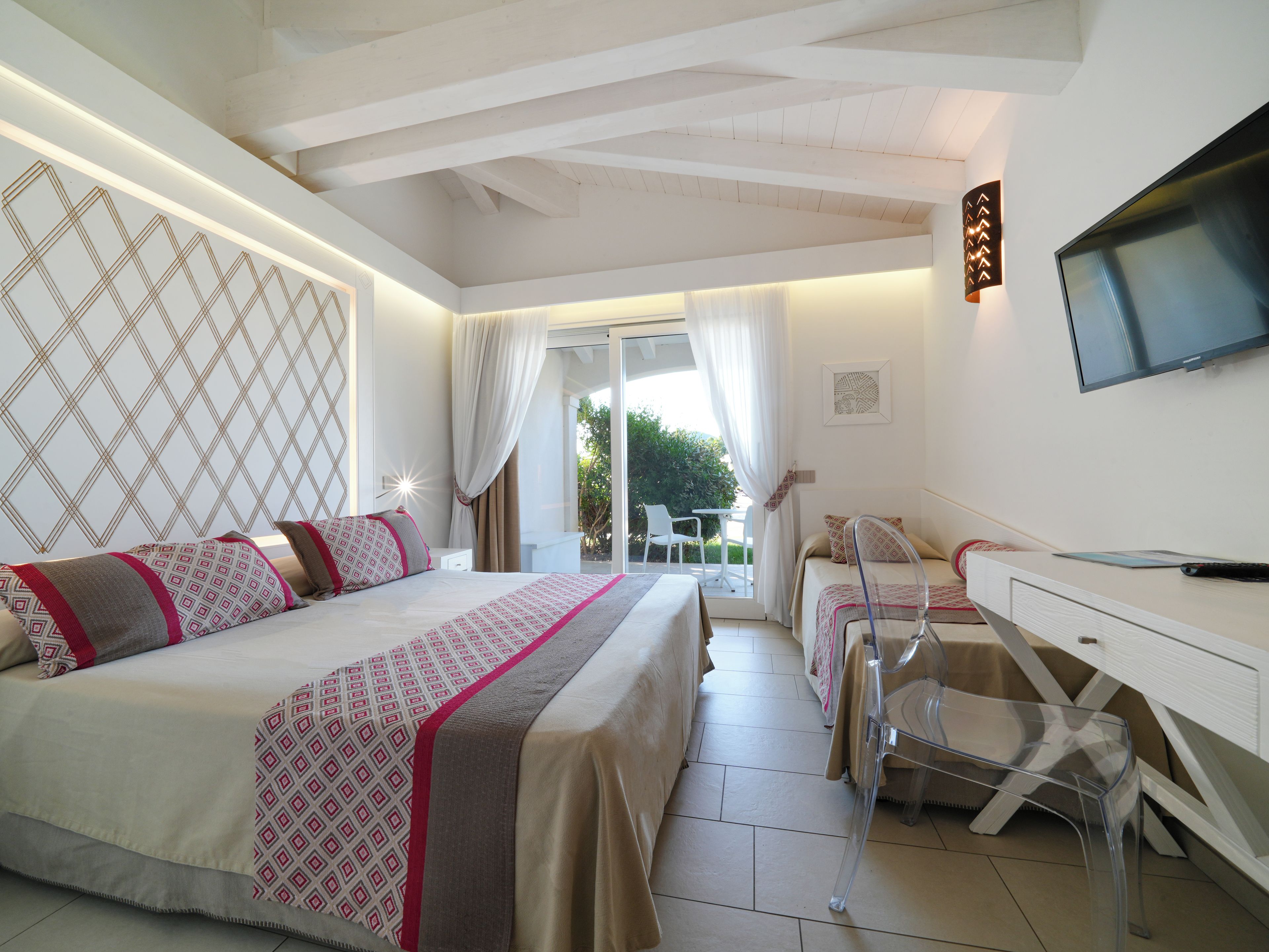 A hotel room with a bed, a desk, a chair, a TV, and a balcony with a view of greenery.