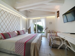 A hotel room with a bed, a desk, a chair, a TV, and a balcony with a view of greenery.