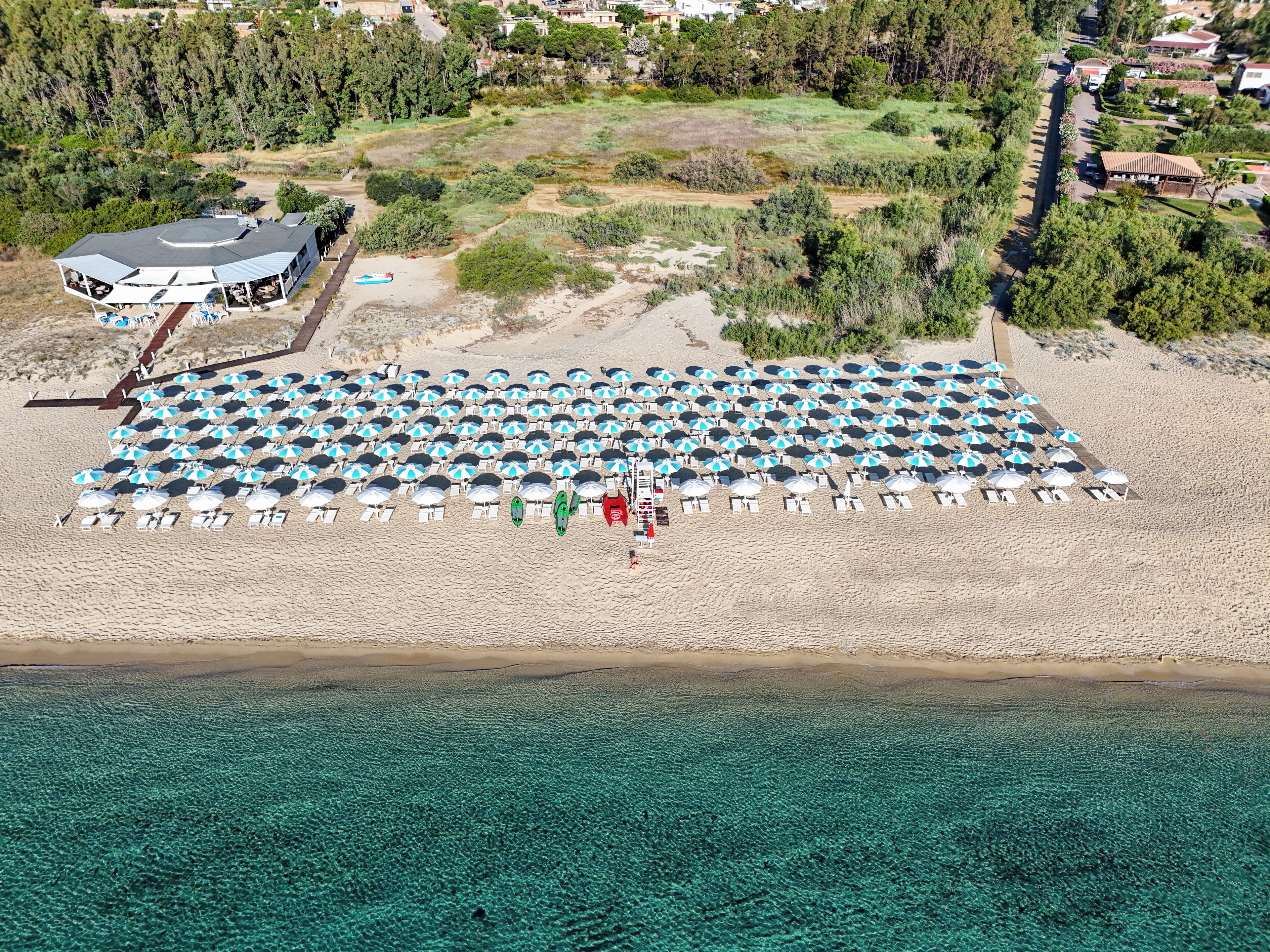 Una vista aerea di una spiaggia con numerosi ombrelloni e lettini disposti in file, un edificio con tetto spiovente e una limpida vista del mare turchese.