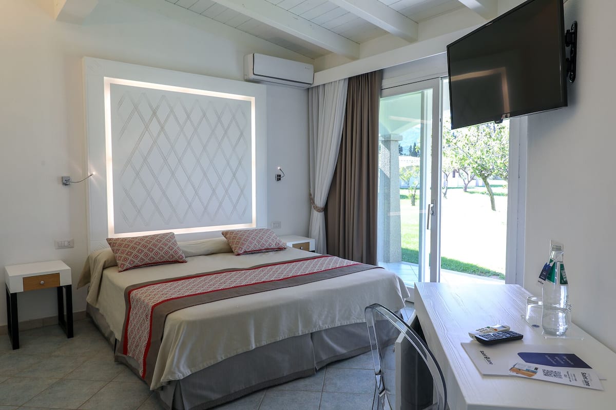 Un dormitorio con cama, escritorio con televisor, ventana con cortinas y unidad de aire acondicionado.