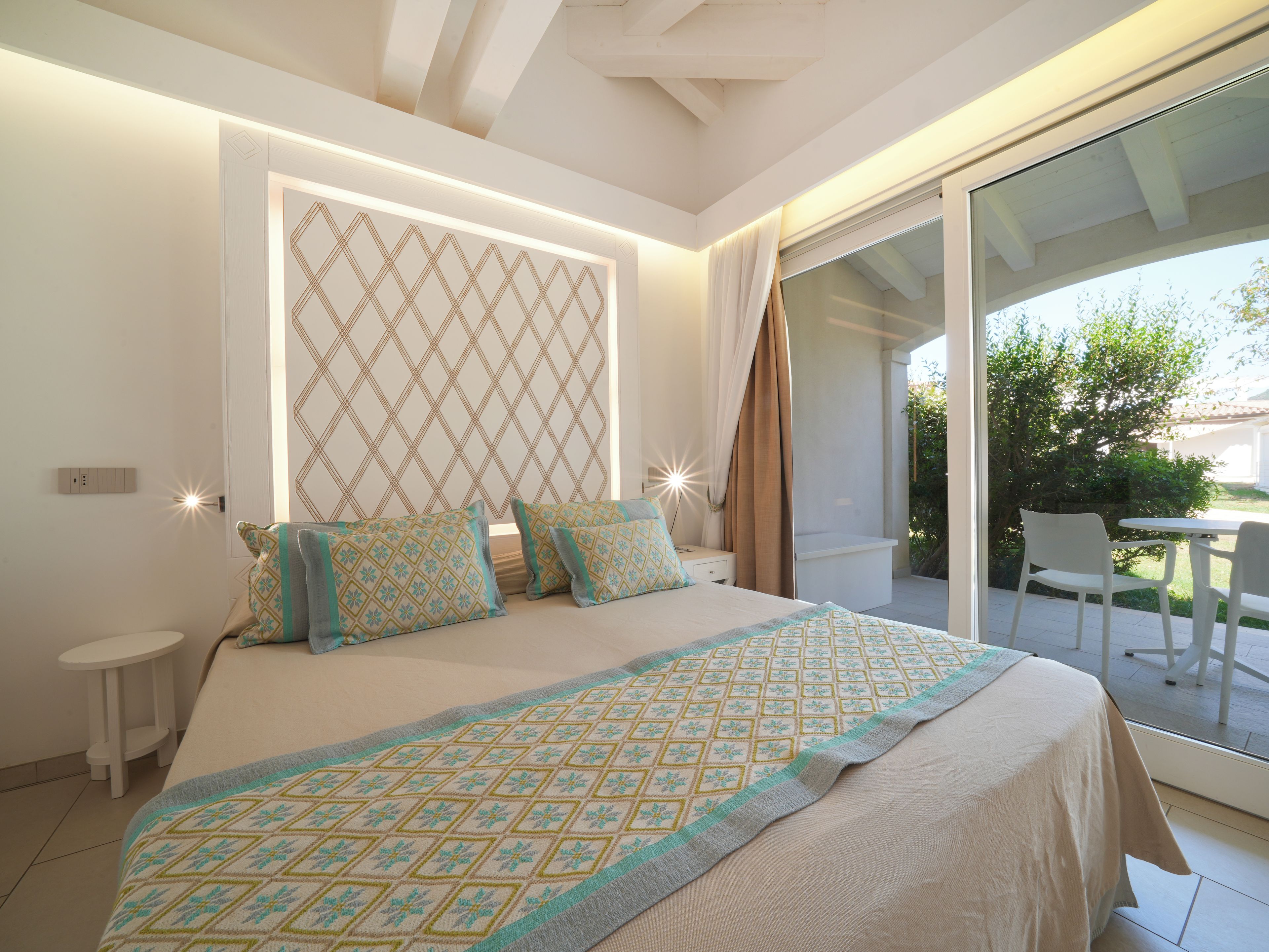 A bedroom with a large bed, a nightstand, a bedside table, and a view of the outdoors through a sliding glass door.