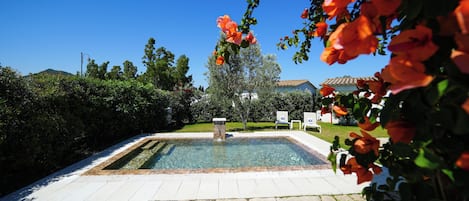A swimming pool with a stone deck, surrounded by greenery and a clear sky.