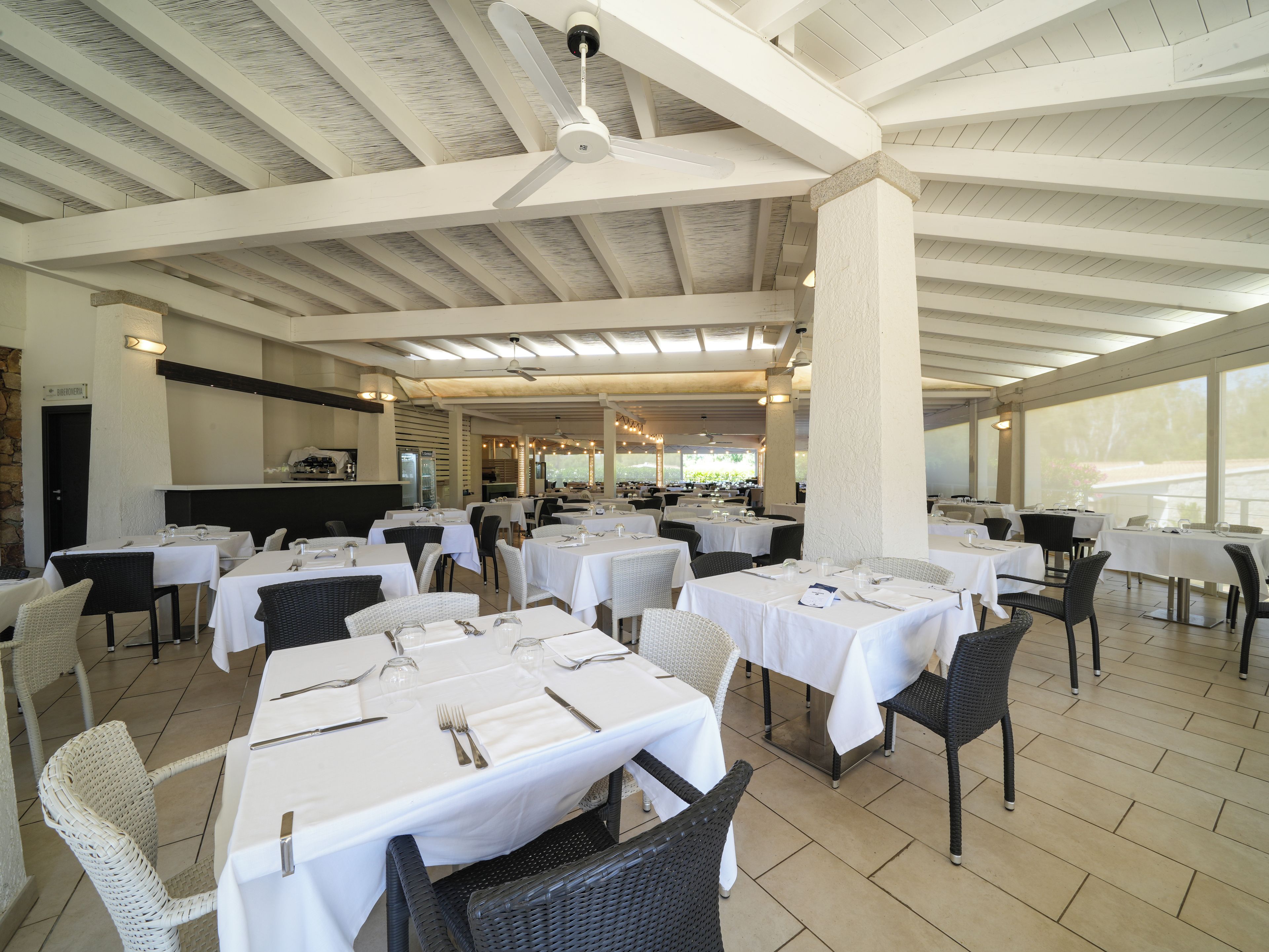 An indoor dining area with tables set for service, a ceiling fan, and a view through large windows.