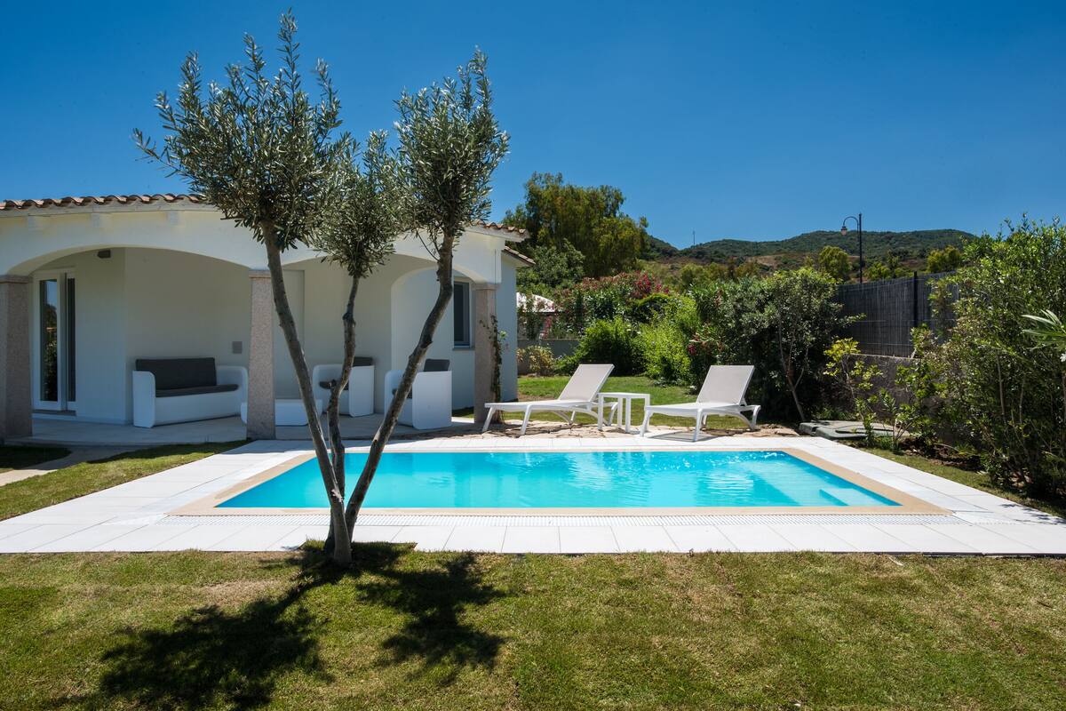 Una piscina con sillas de descanso, un edificio blanco con arcos y un árbol en primer plano.