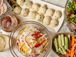 Une table sur laquelle on trouve divers aliments, notamment des légumes coupés en tranches, un bol de légumes râpés, une assiette de boulettes de viande et un verre de jus.