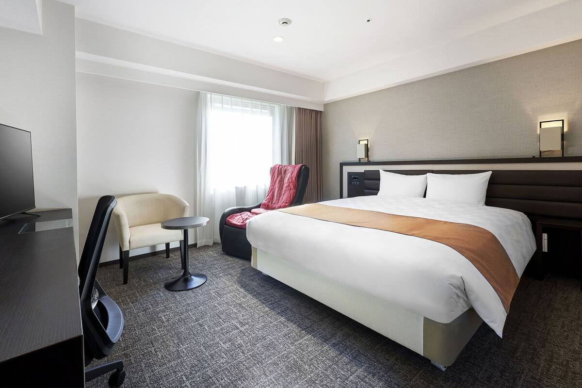A hotel room with a large bed, a desk with a chair, a TV, and a window with curtains.