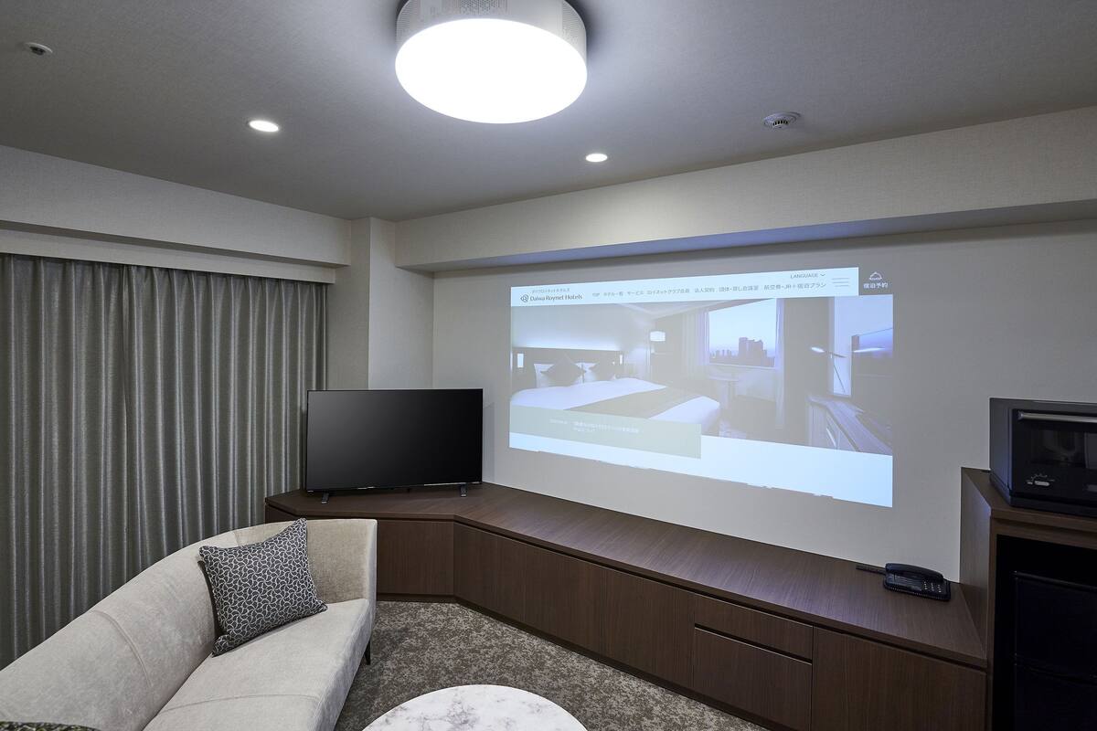 A modern hotel room with a large flat-screen TV, a sofa, a coffee table, and a projection screen displaying a hotel interior.
