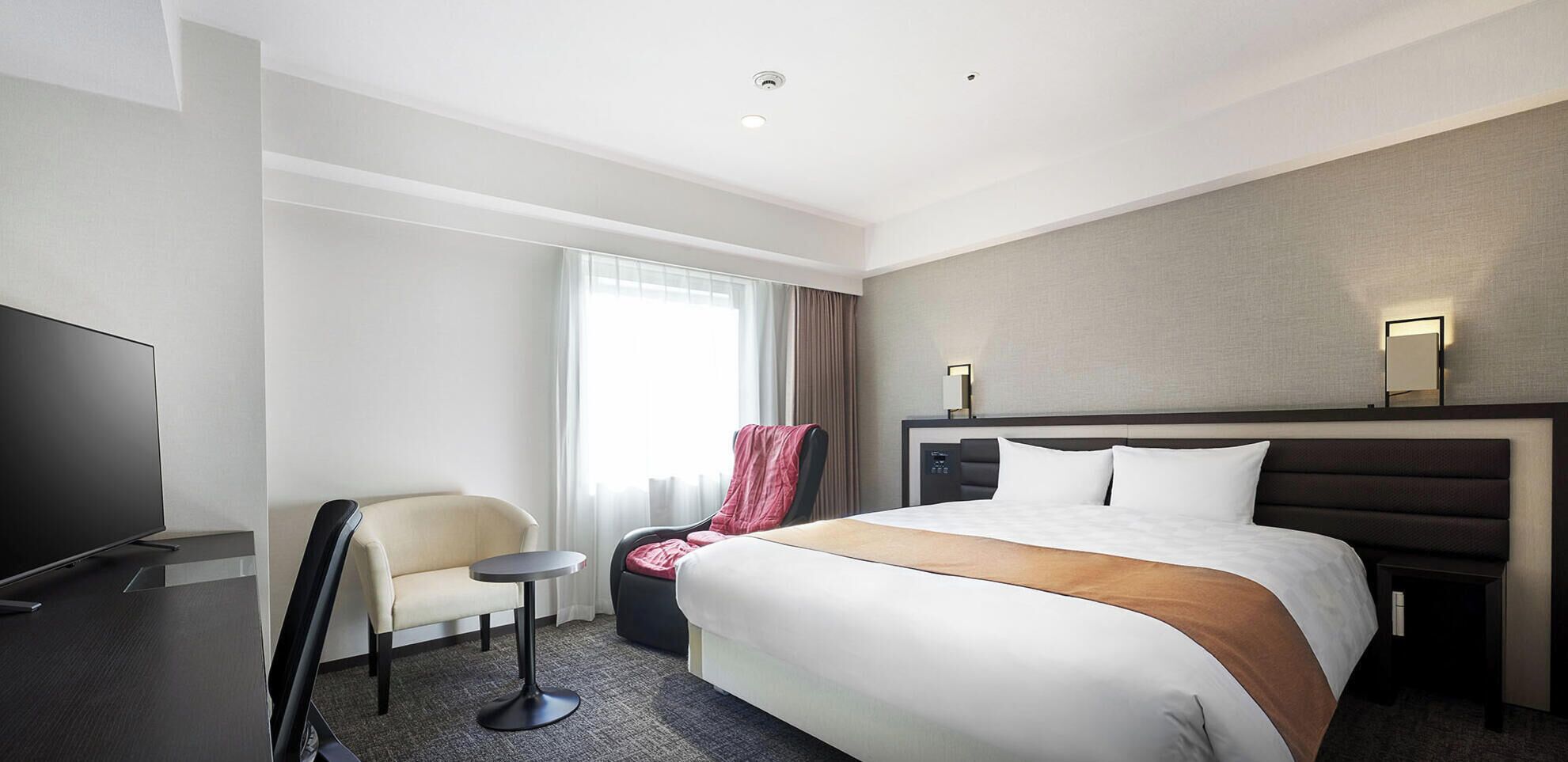 Une chambre d’hôtel avec un lit, une télévision, une chaise, une petite table et une fenêtre avec des rideaux.