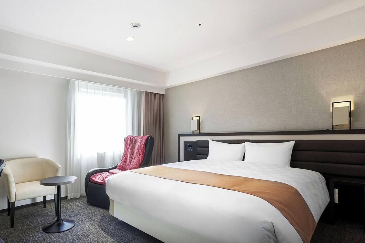 A hotel room with a bed, a TV, a chair, a small table, and a window with curtains.