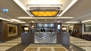 A hotel reception area with a marble counter, two receptionists, a large chandelier, and a framed cityscape artwork on the wall. - Nidya Hotel Galataport (Istanbul)