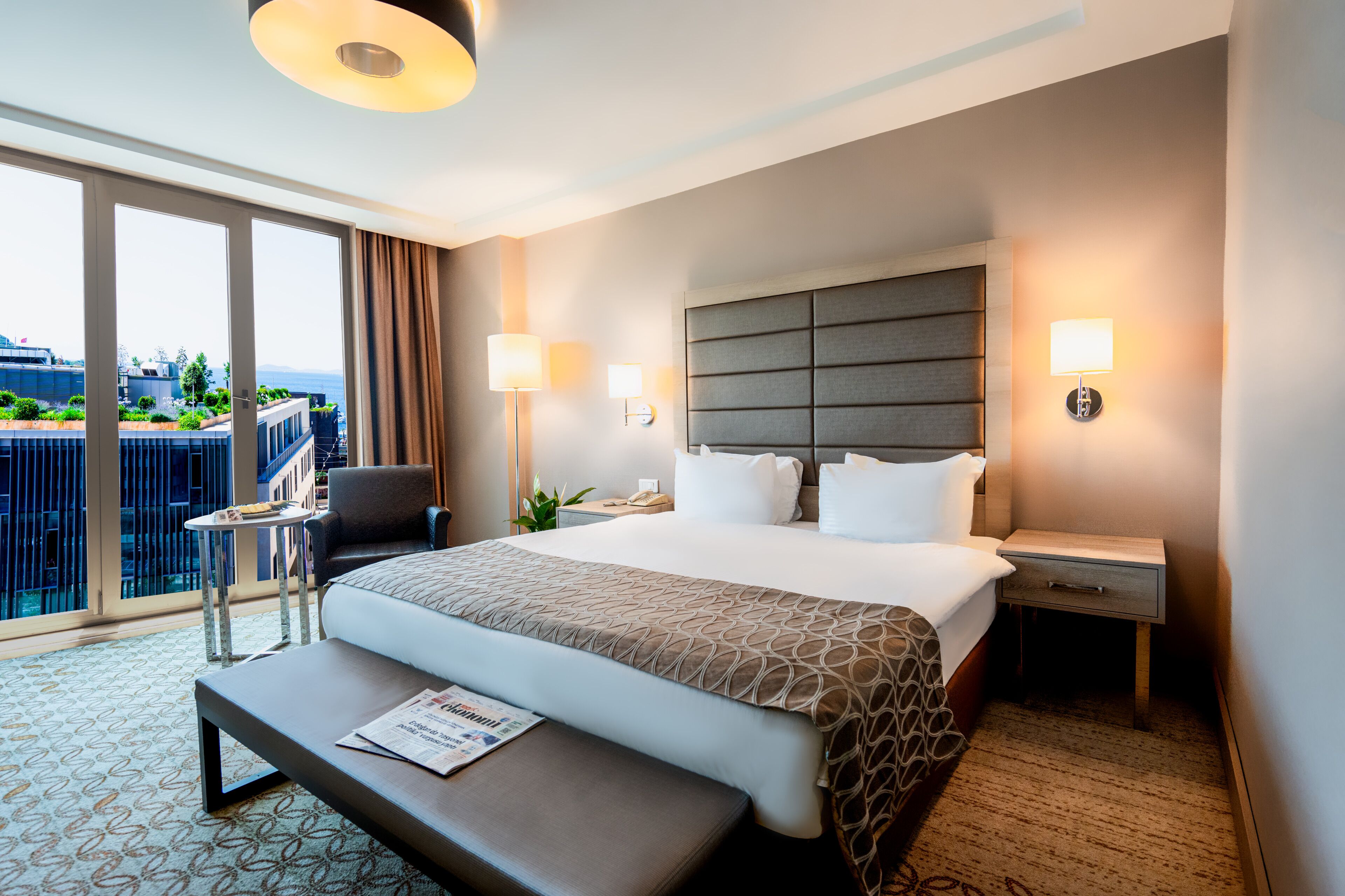 A hotel room with a large bed, a bedside table, a chair, and a view of the city.
