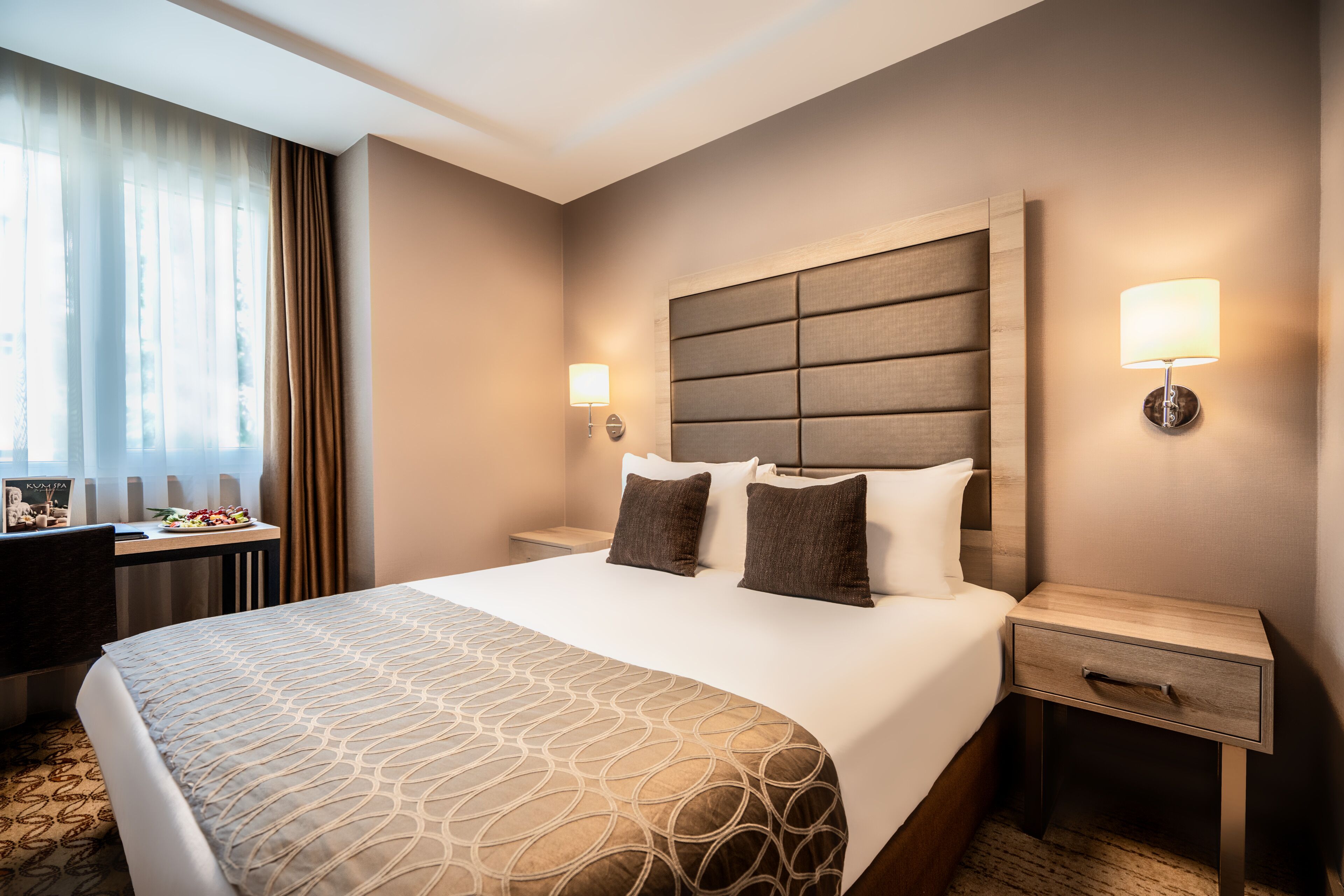 Hotellihuone, jossa on suuri sänky, kaksi yöpöytää, työpöytä ja ikkuna, jossa on verhot.