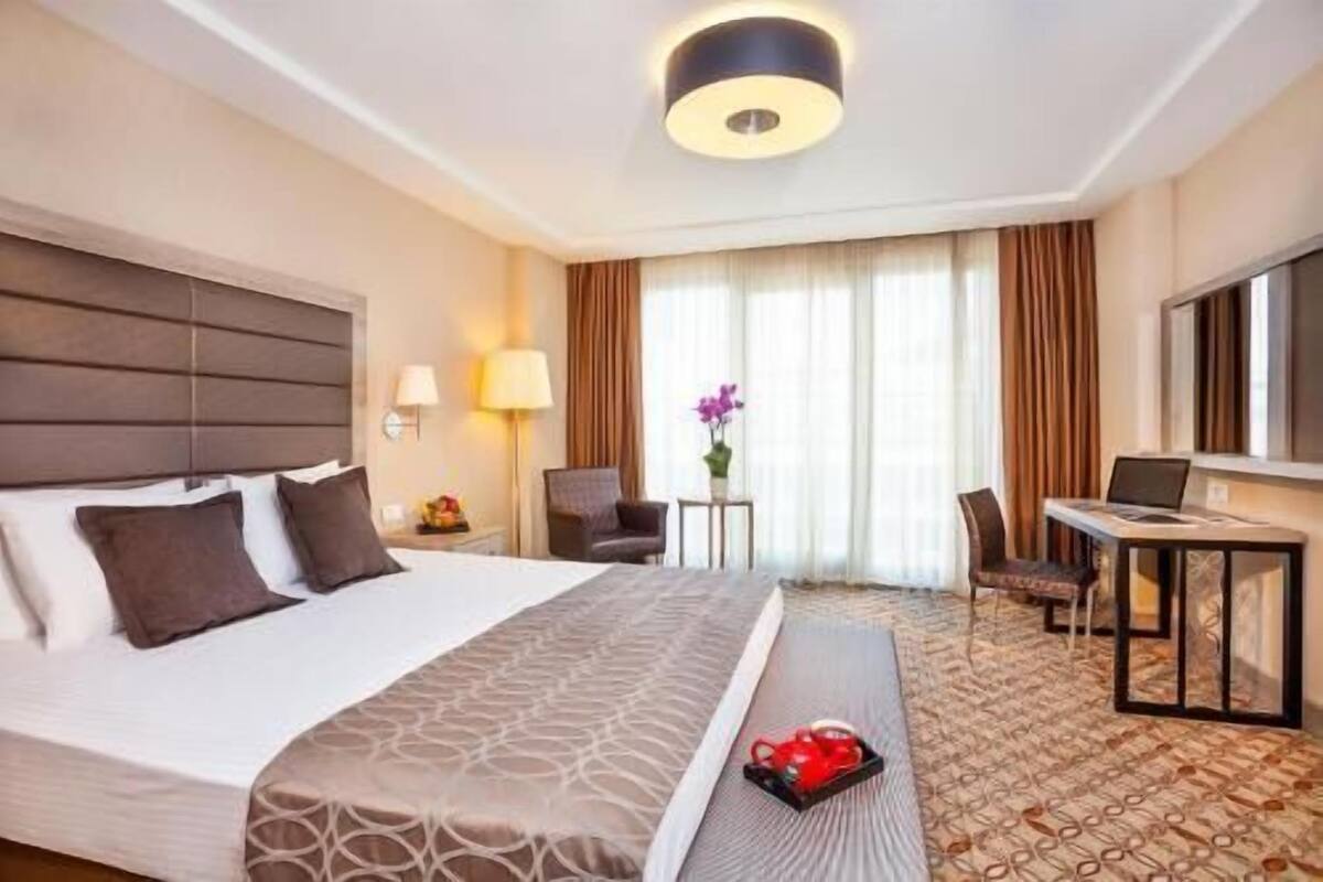 A hotel room with a large bed, a desk, a chair, a lamp, and a window with curtains.