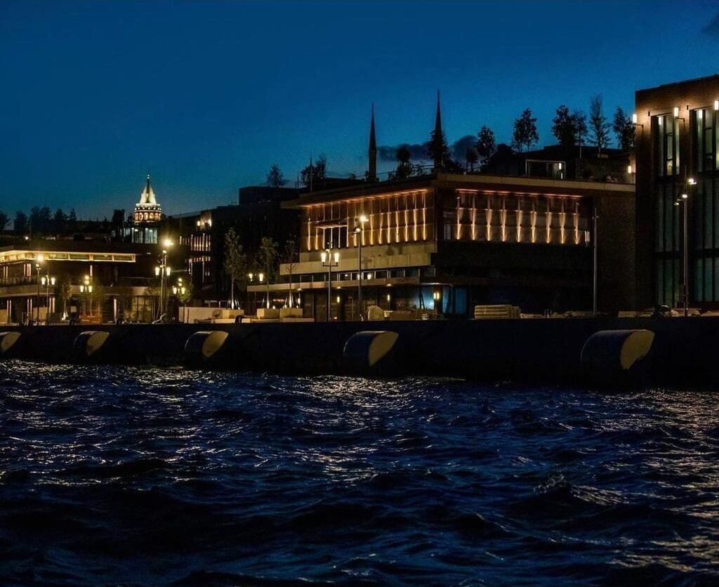 A waterfront view with buildings, including a mosque with a minaret, and a modern building with illuminated windows.
