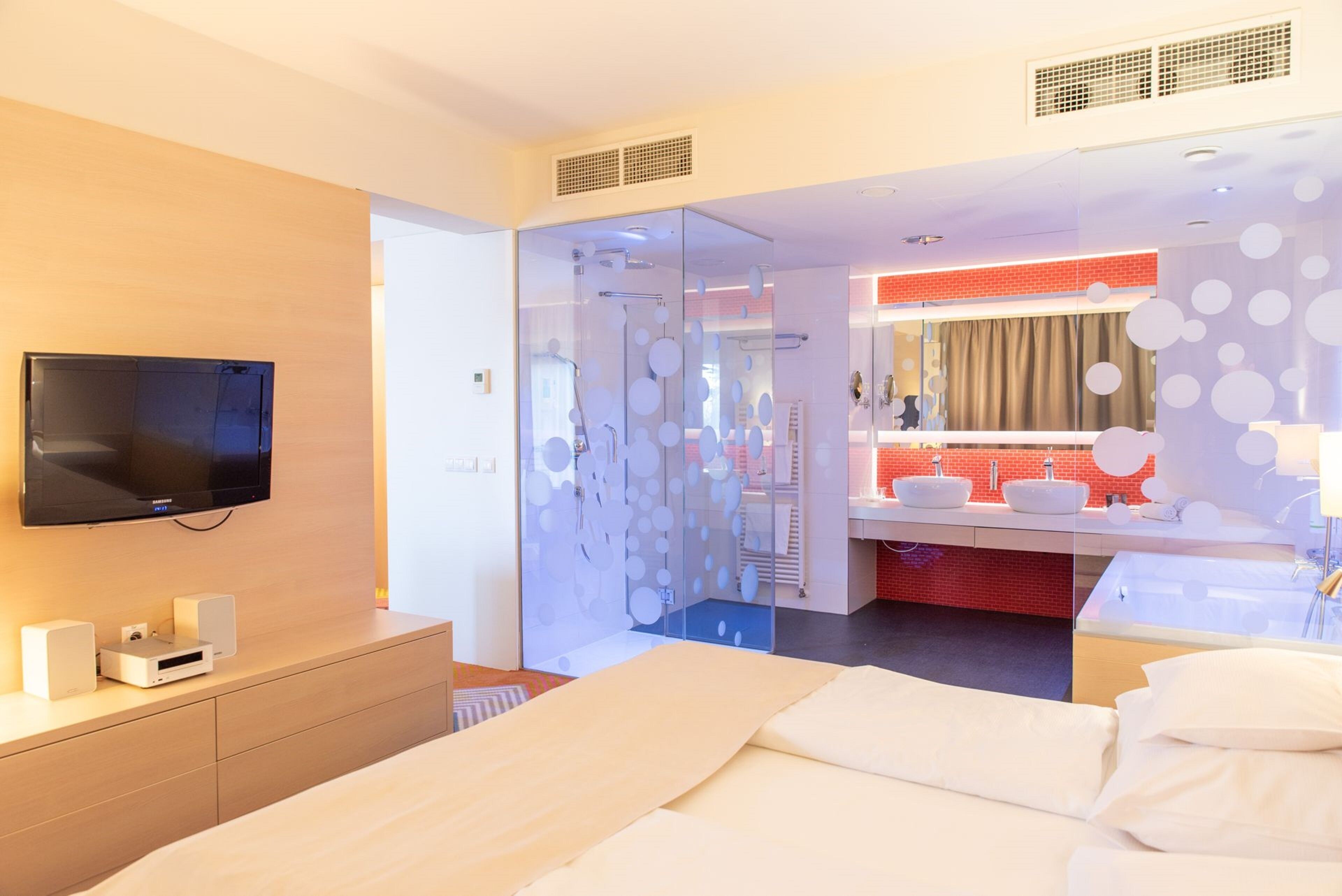 A modern hotel room with a large bed, a TV mounted on the wall, a bathroom with a red-tiled wall, and a glass partition with circular patterns.