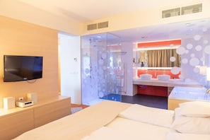 Une chambre d’hôtel moderne dotée d’un grand lit, d’un téléviseur fixé au mur, d’une salle de bain avec un mur recouvert de carreaux rouges et d’une cloison en verre ornée de motifs circulaires.