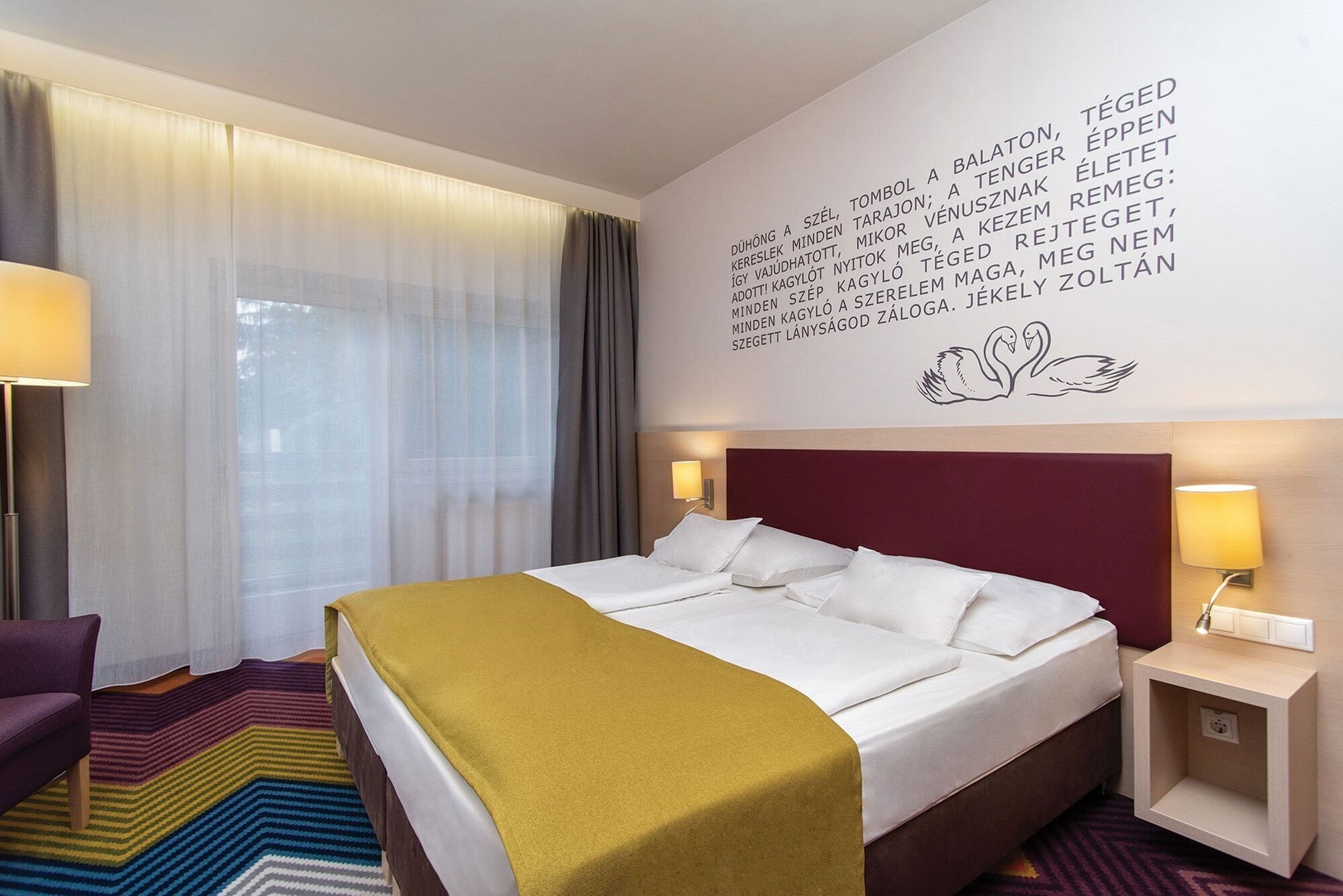 Ein Hotelzimmer mit einem Bett, einem Sessel, einem Nachttisch und einer Wand mit Text und einer Schwanillustration.