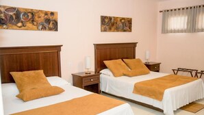 A hotel room with two beds, each with a wooden headboard, white bedding, and a brown throw blanket. There are two bedside tables with lamps, a window with curtains, and abstract wall art. - Allamanda Hotel (Petionville)