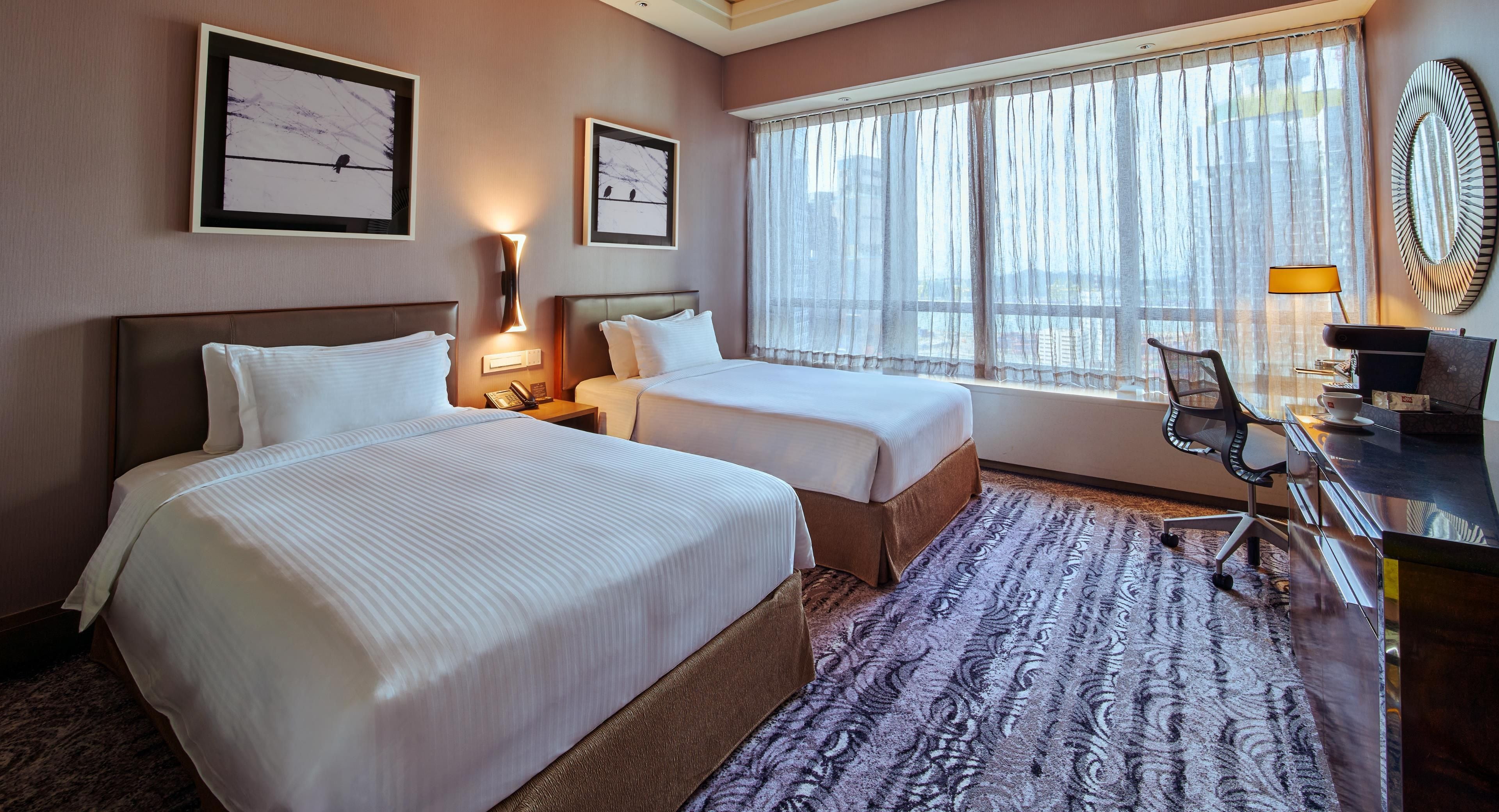 A hotel room with two beds, a desk with a chair, a window with curtains, and framed artwork on the wall.