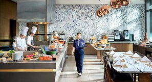 Una cocina moderna con chefs preparando alimentos, un bufé con diversos platos y una pared de mármol.