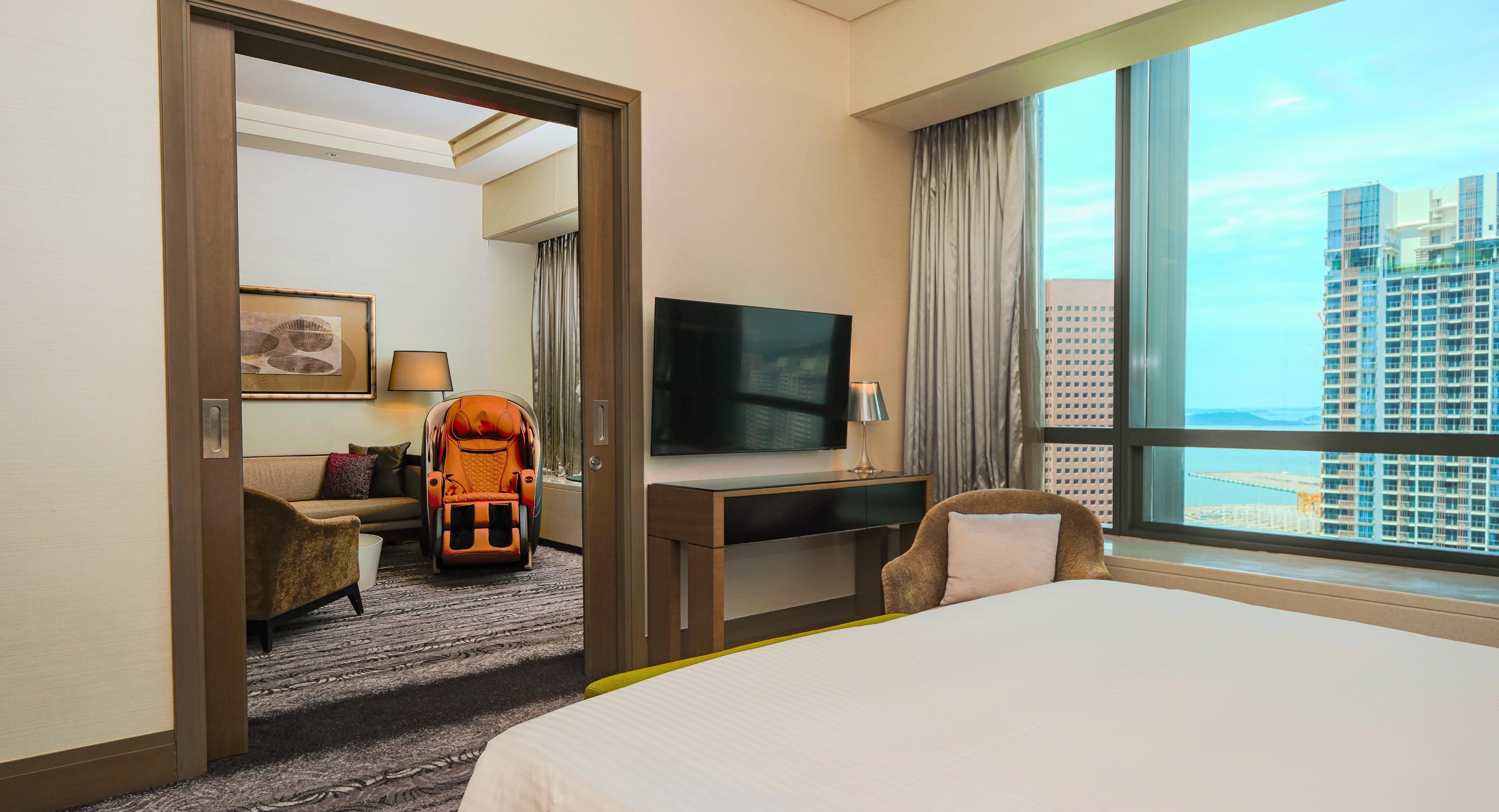 Een hotelkamer met een bed, een bureau met een televisie, een massagestoel en uitzicht op een stadsgezicht.