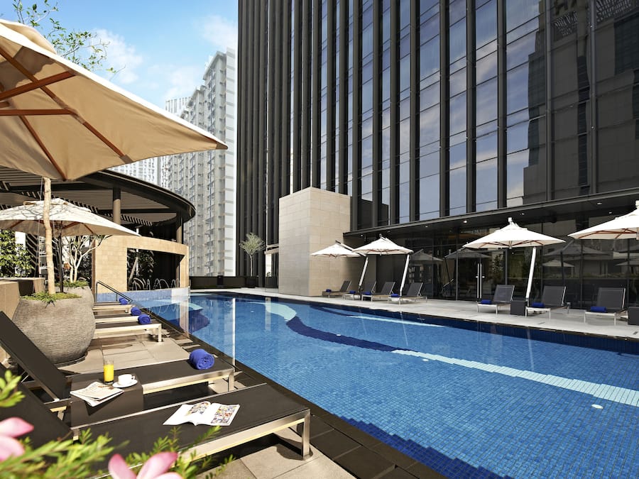 Un área de piscina en la azotea con sillones de descanso y sombrillas, rodeada de edificios altos.