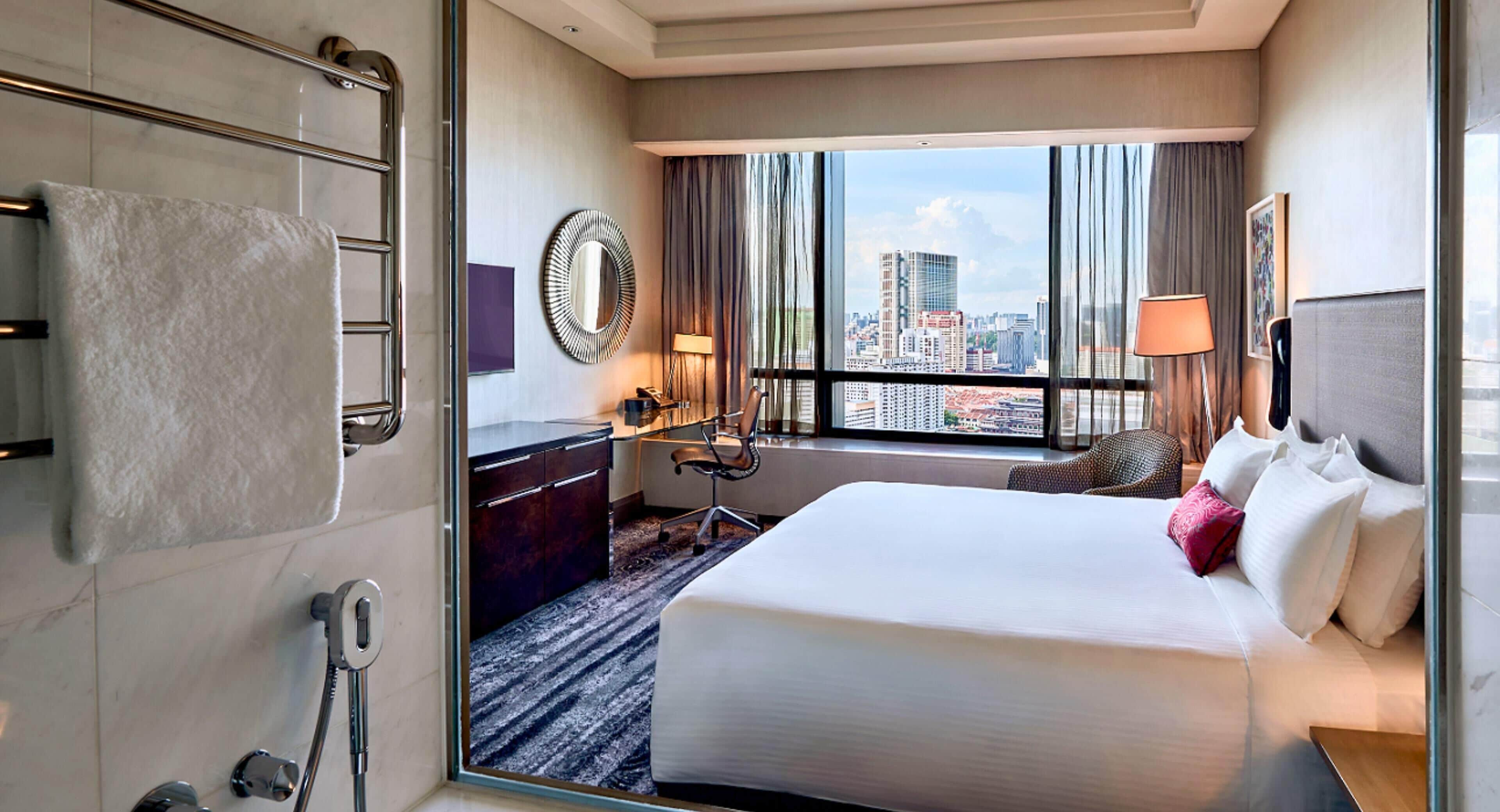 Habitación de hotel con una cama grande, un escritorio, una silla, un baño con toallero y vistas a la ciudad desde la ventana.