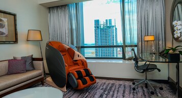 A hotel room with a massage chair, a sofa, a desk with a chair, a window with a view of a building, and a potted plant.